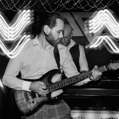 Muzyk gra na gitarze elektrycznej, a za nim stoi drugi mężczyzna. Efekt fal świetlnych na zdjęciu to zasługa długiego czasu naswietlania.