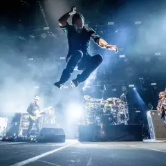 Energiczna fotografia koncertowa: wokalista w powietrzu, gitarzyści i perkusista na scenie.