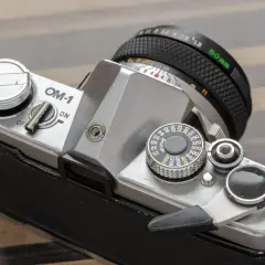 Srebrny aparat OM-1 z obiektywem 50mm. Widoczny pokrętło czułości ISO i dźwignia naciągu migawki.