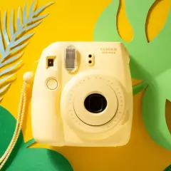 Żółty aparat Fujifilm Instax Mini 8 z paskiem, otoczony zielonymi liśćmi monstery i niebieskimi liśćmi paproci. Idealny do zdjęć z **samowyzwalaczem**.