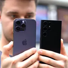 Mężczyzna trzyma dwa najnowsze smartfony: fioletowy iPhone i czarny Samsung. Oba mają imponujące aparaty, gotowe do robienia świetnych zdjęć.