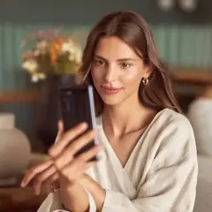 Kobieta robi sobie selfie w lustrze, trzymając telefon. W tle wazon z kwiatami i meble.