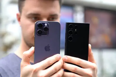Mężczyzna trzyma dwa najnowsze smartfony: fioletowy iPhone i czarny Samsung. Oba mają imponujące aparaty, gotowe do robienia świetnych zdjęć.
