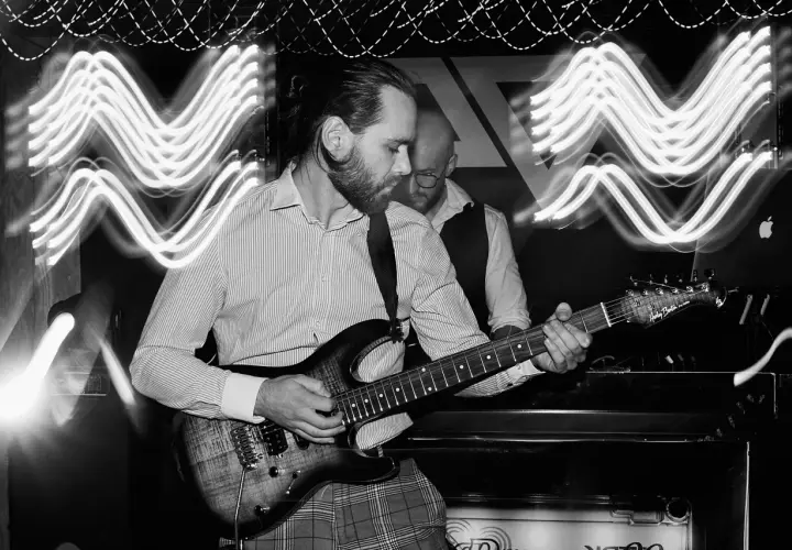 Muzyk gra na gitarze elektrycznej, a za nim stoi drugi mężczyzna. Efekt fal świetlnych na zdjęciu to zasługa długiego czasu naswietlania.