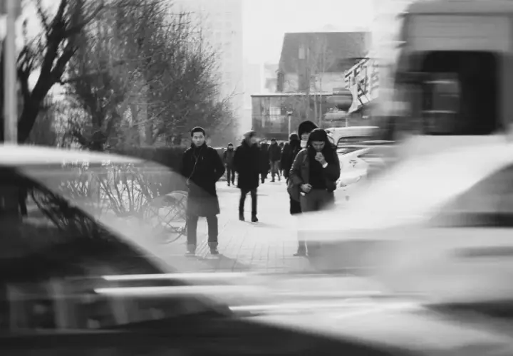 Ludzie na chodniku, rozmyte samochody i drzewa. Długi czas naświetlania (shutter speed) uchwycił ruch uliczny i miejską atmosferę.