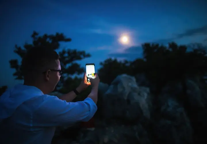 Chłopak z telefonem Samsung próbuje zrobić zdjęcie księżyca. Nocne niebo, drzewa i skały tworzą tło.