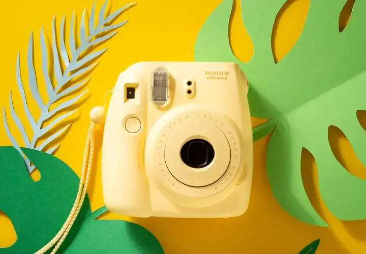 Żółty aparat Fujifilm Instax Mini 8 z paskiem, otoczony zielonymi liśćmi monstery i niebieskimi liśćmi paproci. Idealny do zdjęć z **samowyzwalaczem**.