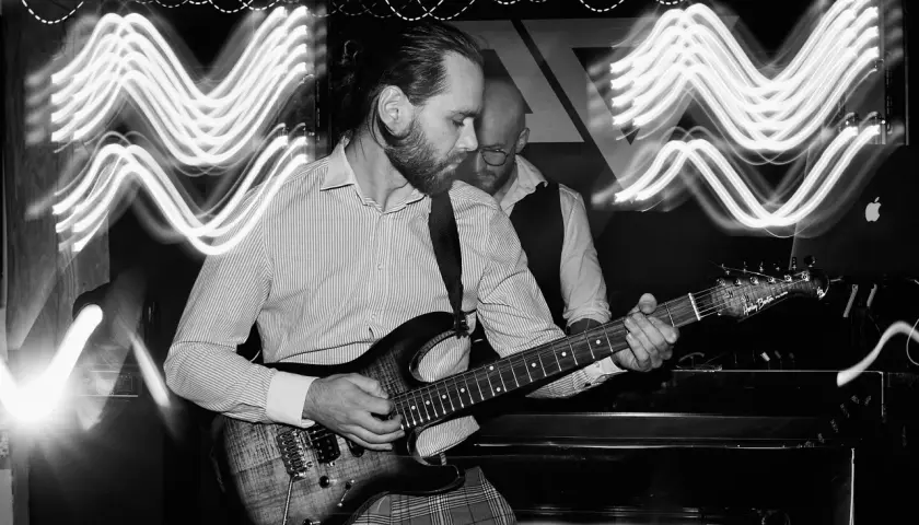 Muzyk gra na gitarze elektrycznej, a za nim stoi drugi mężczyzna. Efekt fal świetlnych na zdjęciu to zasługa długiego czasu naswietlania.