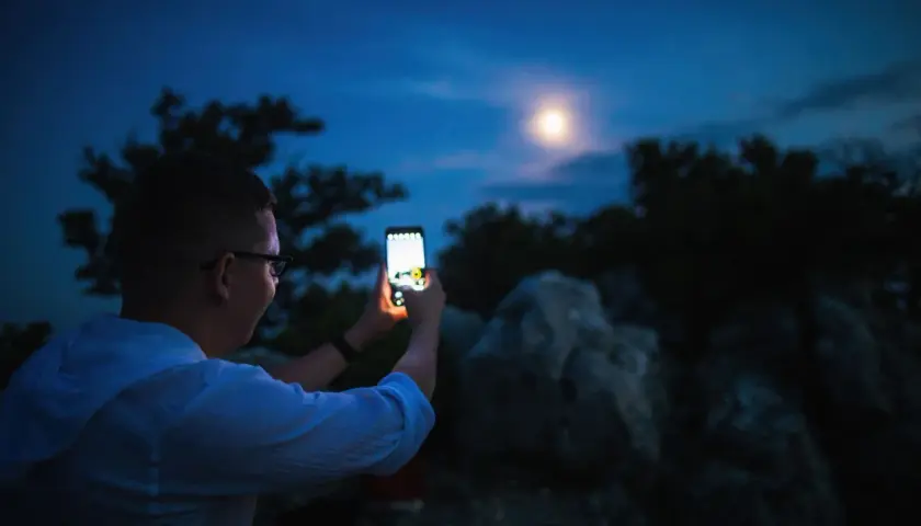 Chłopak z telefonem Samsung próbuje zrobić zdjęcie księżyca. Nocne niebo, drzewa i skały tworzą tło.