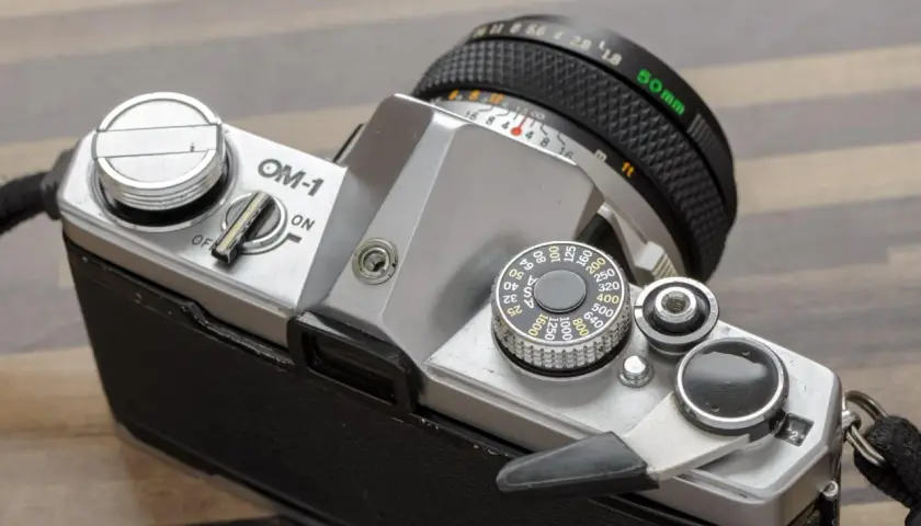 Srebrny aparat OM-1 z obiektywem 50mm. Widoczny pokrętło czułości ISO i dźwignia naciągu migawki.