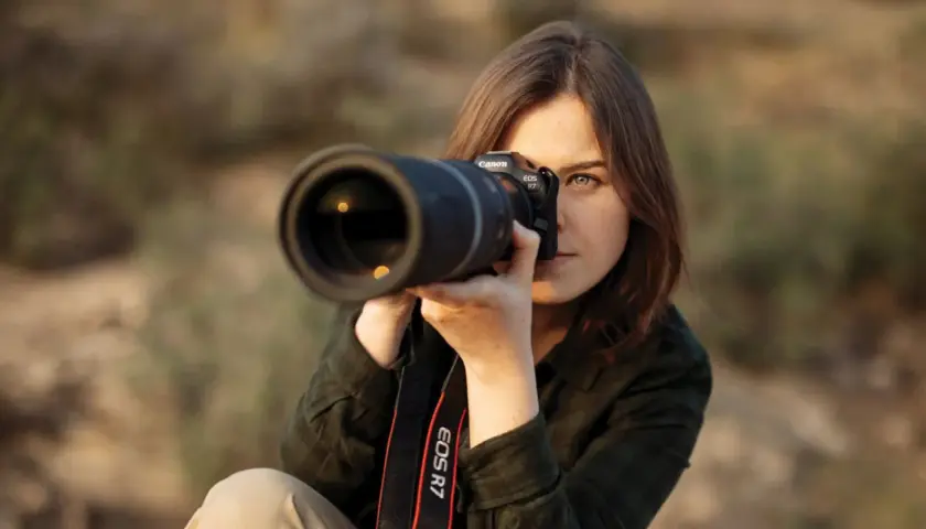Kobieta z aparatem Canon EOS R7, skupiona na kadrze. Dopasowuje ustawienia aparatu, by uchwycić idealny moment.
