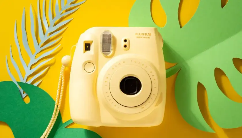 Żółty aparat Fujifilm Instax Mini 8 z paskiem, otoczony zielonymi liśćmi monstery i niebieskimi liśćmi paproci. Idealny do zdjęć z **samowyzwalaczem**.