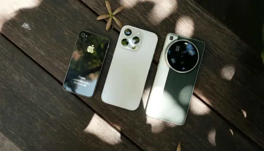 Trzy smartfony fotograficzne: stary iPhone, biały iPhone i zielony Huawei z aparatem Leica, leżą na drewnianym stole.