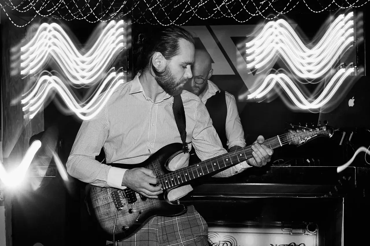 Muzyk gra na gitarze elektrycznej, a za nim stoi drugi mężczyzna. Efekt fal świetlnych na zdjęciu to zasługa długiego czasu naswietlania.