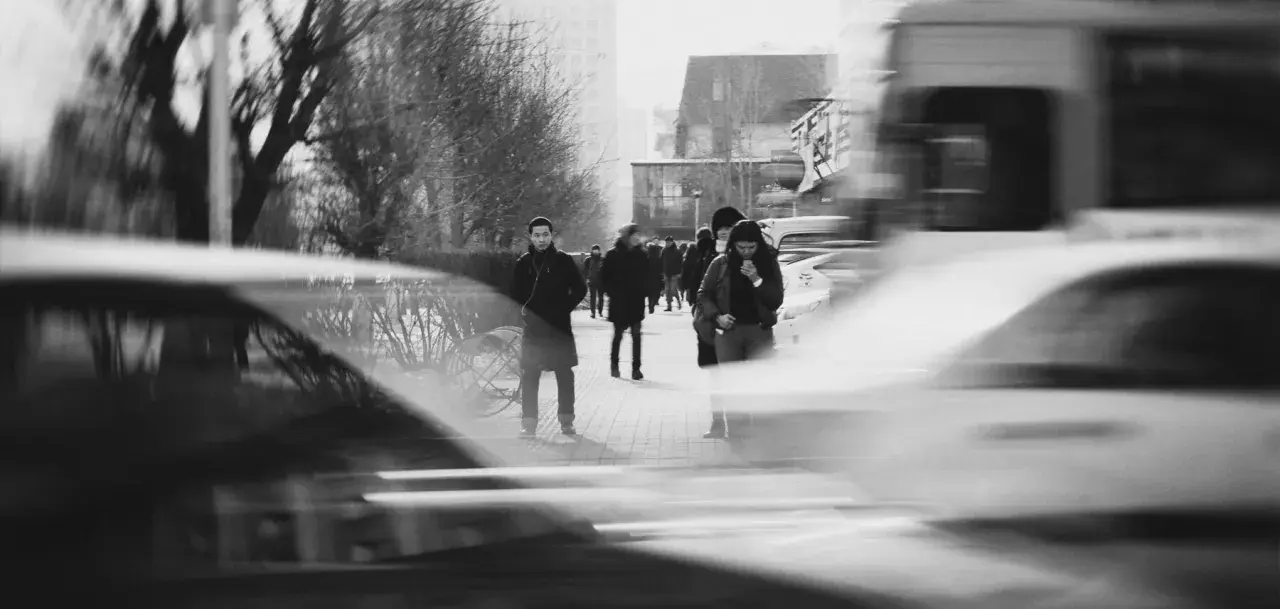 Ludzie na chodniku, rozmyte samochody i drzewa. Długi czas naświetlania (shutter speed) uchwycił ruch uliczny i miejską atmosferę.