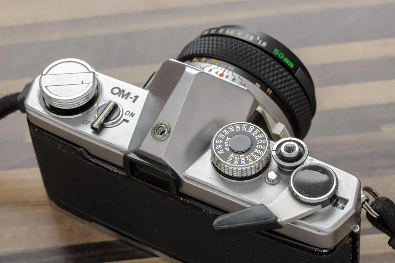 Srebrny aparat OM-1 z obiektywem 50mm. Widoczny pokrętło czułości ISO i dźwignia naciągu migawki.