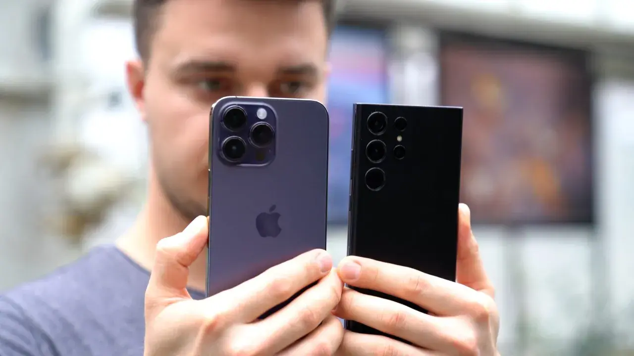 Mężczyzna trzyma dwa najnowsze smartfony: fioletowy iPhone i czarny Samsung. Oba mają imponujące aparaty, gotowe do robienia świetnych zdjęć.